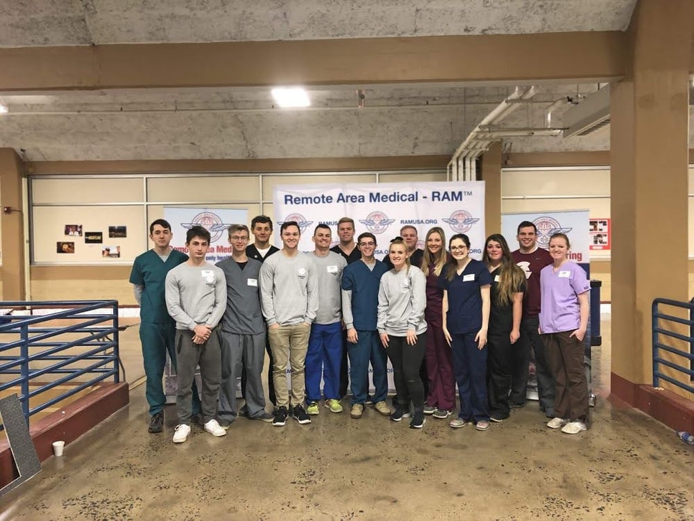 A group of thirteen people in scrubs and casual clothes, including aspiring healthcare professionals, stand indoors in front of a Remote Area Medical - RAM banner, posing for a group photo.