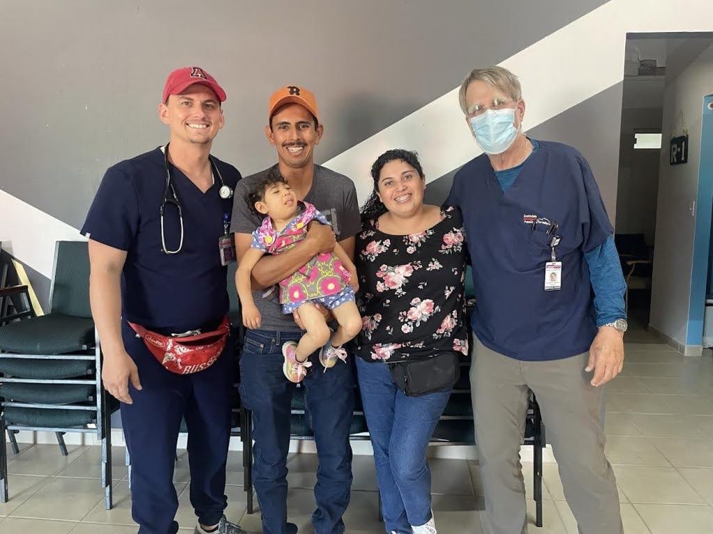 Two healthcare professionals stand with a man holding a young girl and a woman, all smiling indoors in a waiting room setting.