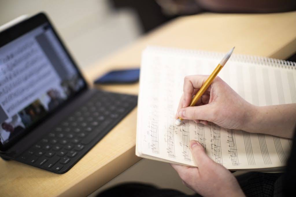 Person writing music notes on sheet paper with a pencil, while referencing a tablet displaying sheet music on a desk.